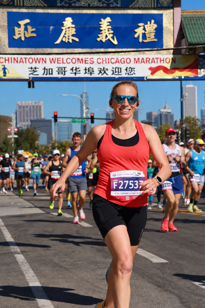 A female runner in a red tank top and black shorts smiles while running in the Chicago Marathon, with a Chinatown welcome banner in the background and other runners nearby.