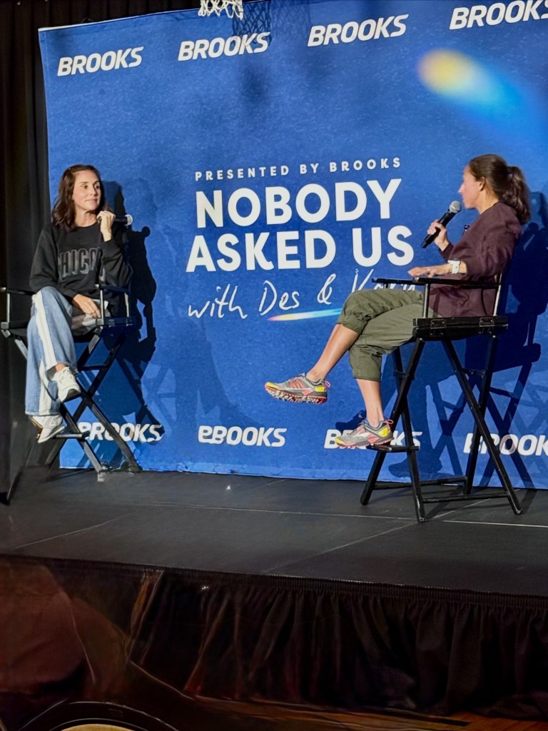 Two women seated on high chairs during a live podcast event titled 'Nobody Asked Us' presented by Brooks. They are engaging in conversation, with a backdrop featuring the Brooks logo.