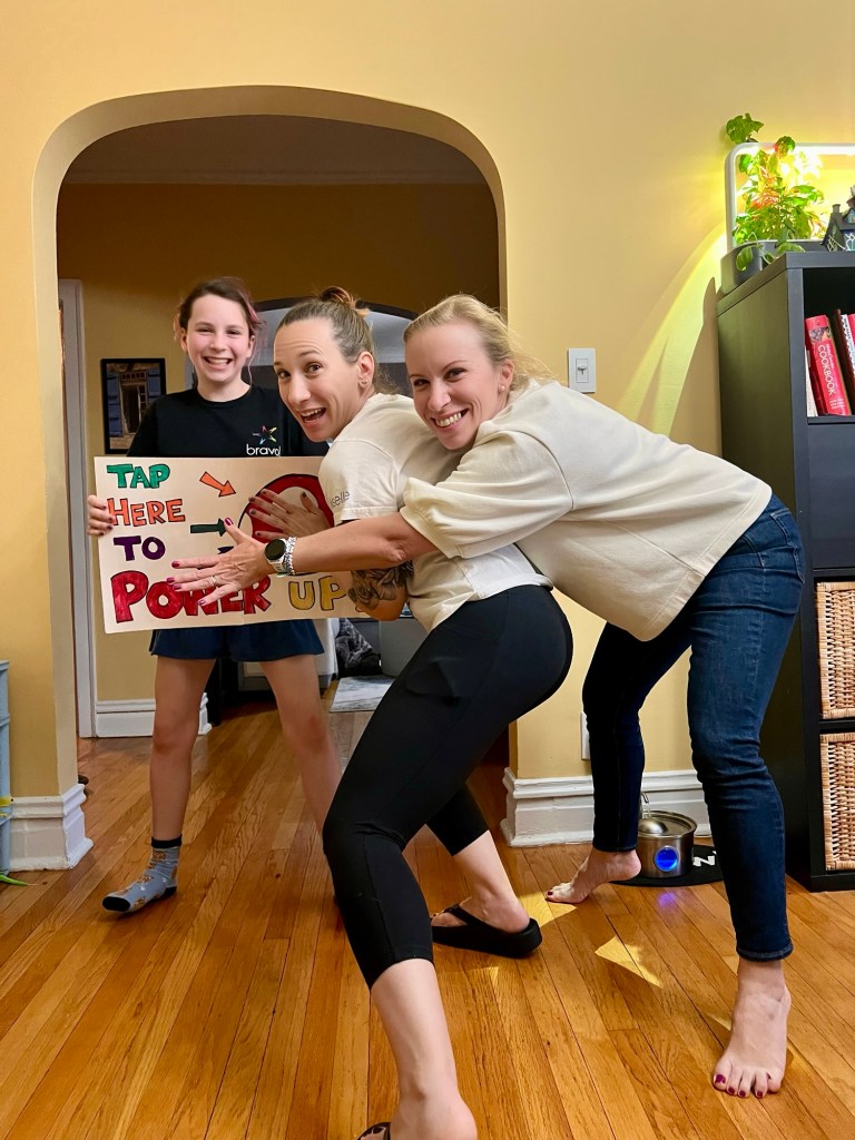 A group of three people in a home setting, with a girl holding a colorful sign that says 'TAP HERE TO POWER UP' while smiling. Two women are playfully posing behind her, one with arms around the other, creating a lively and cheerful atmosphere.
