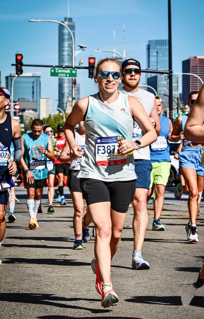 A group of runners participating in a marathon on a city street, with a focus on a woman in sports attire smiling and running confidently. She is wearing sunglasses and a bib number.