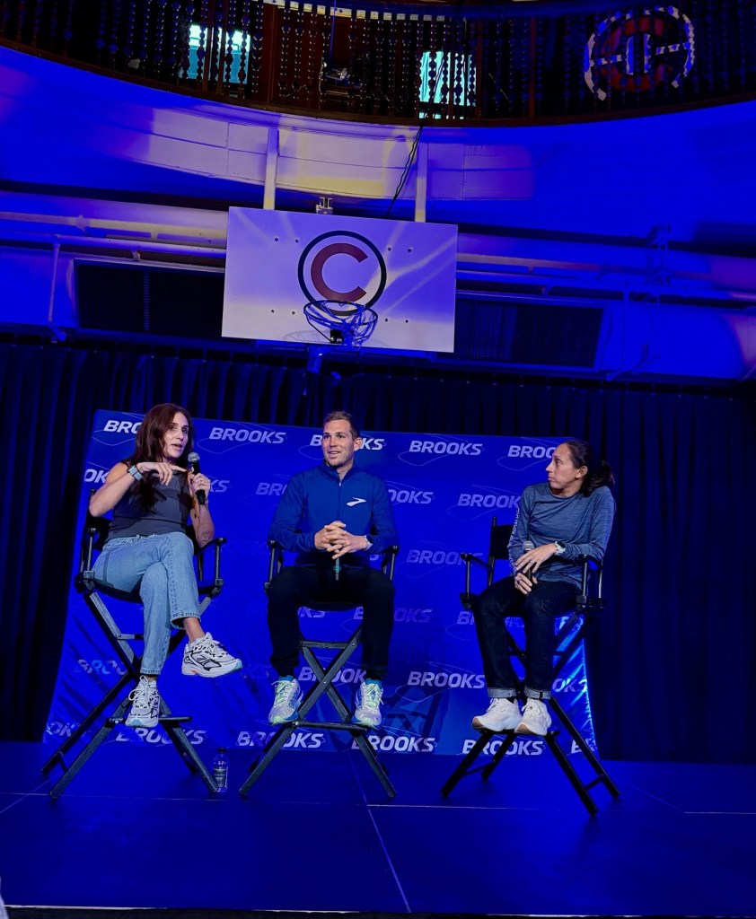 A panel discussion featuring two seated speakers onstage, with one speaker gesturing while holding a microphone. The backdrop displays the Brooks logo against a blue theme.
