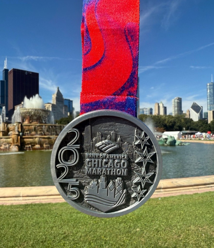 A silver medal for the 2025 Bank of America Chicago Marathon, featuring an engraving of the Chicago skyline and artistic details, hanging in front of a fountain and skyscrapers.