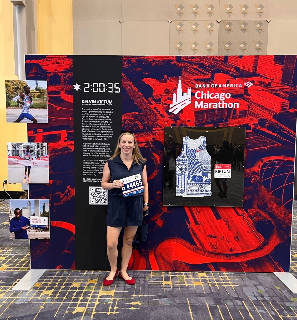 A woman stands in front of a display at the Bank of America Chicago Marathon event, holding her race bib numbered 44465. The backdrop features information about marathon runner Kelvin Kiptum, including his record time of 2:00:35 and a depiction of his racing shirt.