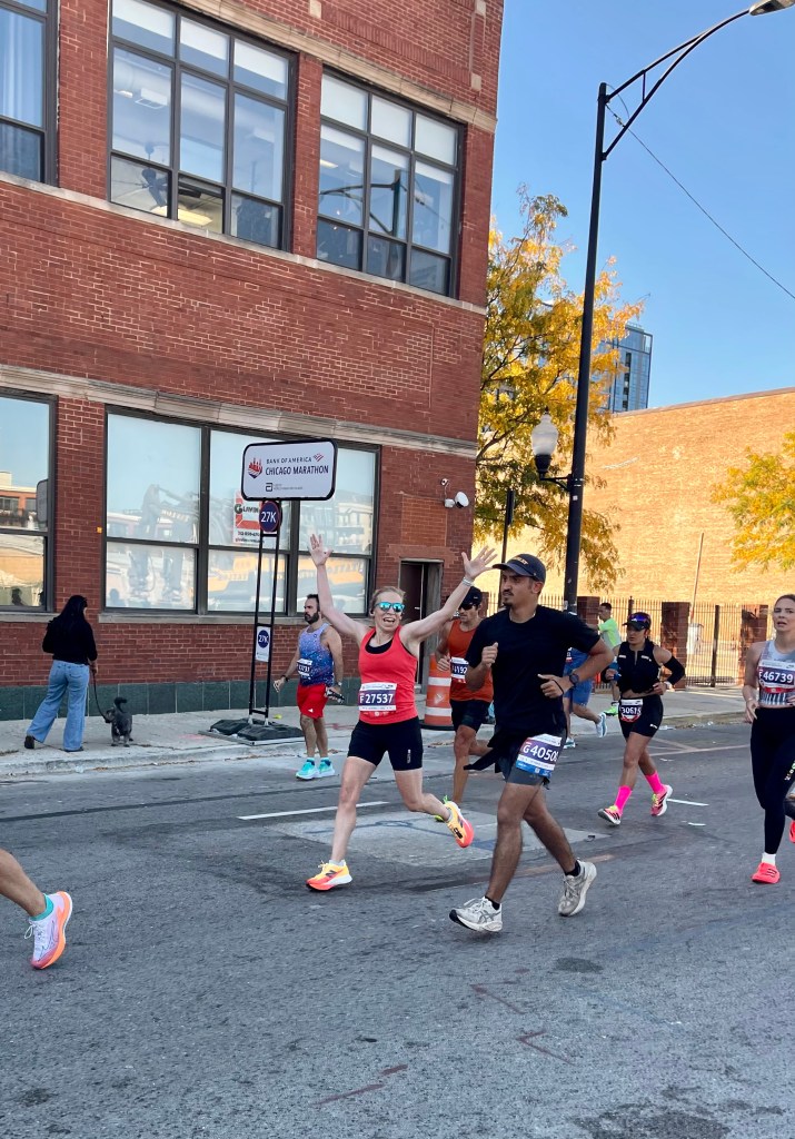 Runners participating in the Chicago Marathon, with a female runner in a red tank top joyfully raising her arms. The scene features a mix of participants and a brick building in the background.