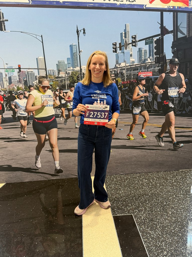 A woman in a blue long-sleeve shirt holding a marathon bib stands in front of a backdrop depicting a city marathon scene with runners.