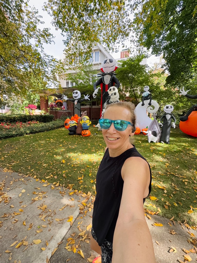 A person taking a selfie in front of a colorful Halloween display featuring inflatable characters, including Jack Skellington and a Minion, surrounded by autumn leaves.