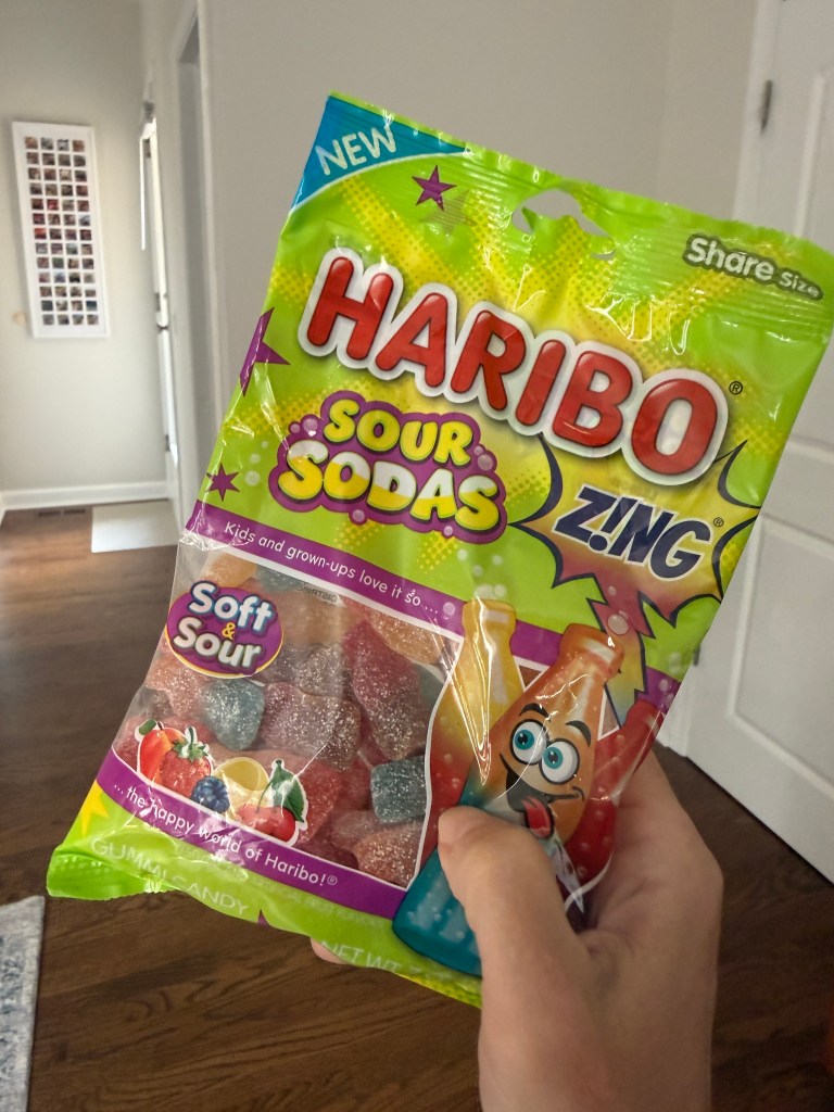 A person holding a bag of Haribo Sour Sodas gummy candy, featuring colorful graphics and the text 'New' and 'Soft & Sour'.