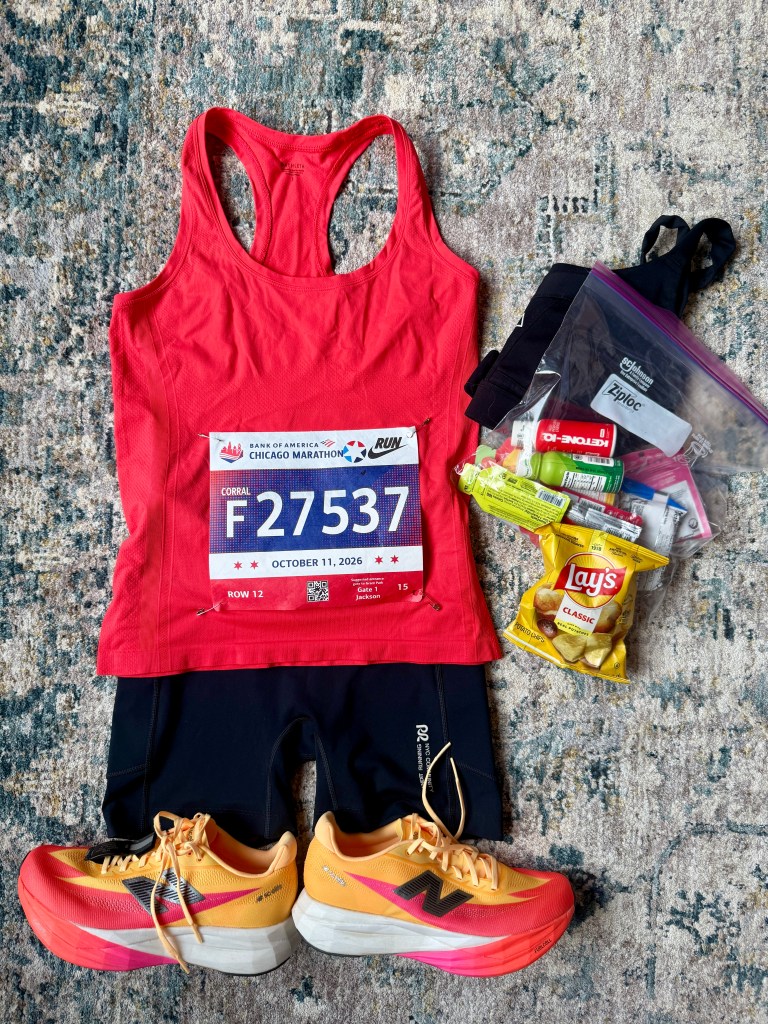 A red tank top with a marathon bib labeled 'F27537', black running shorts, and yellow and pink running shoes, arranged on a patterned rug. Nearby are snacks including Lay's chips and energy gels in a ziplock bag.