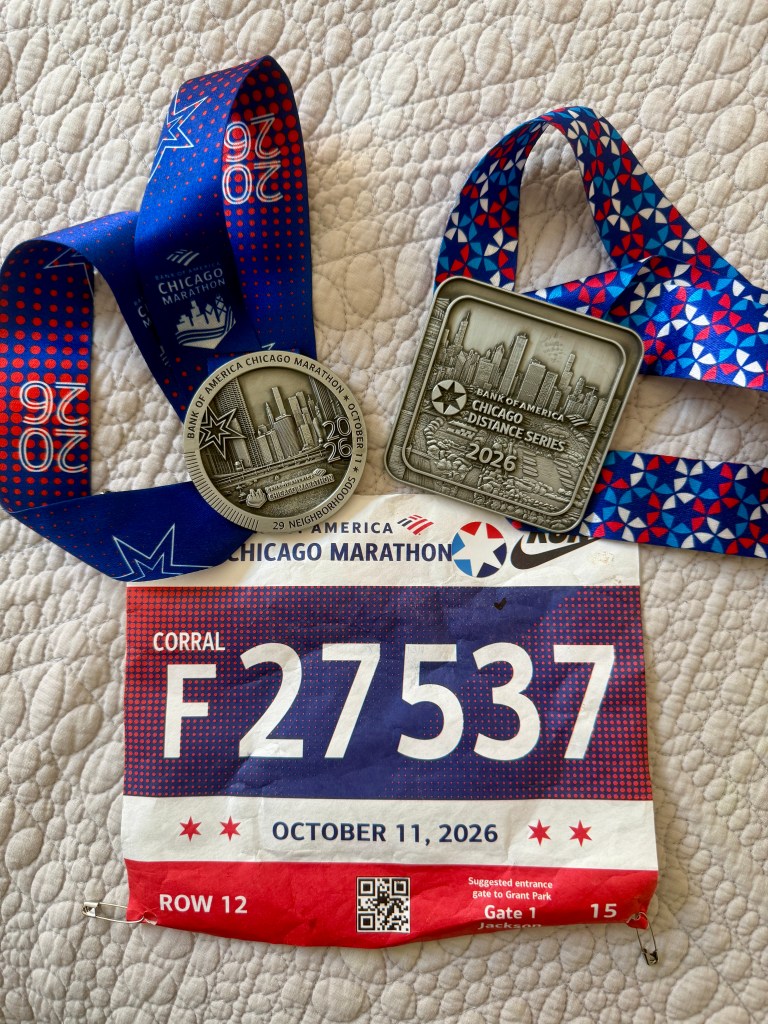 A close-up view of a Chicago Marathon race bib with the number F27537, dated October 11, 2026, alongside two silver medals from the Chicago Marathon and Distance Series, both attached to colorful ribbons.