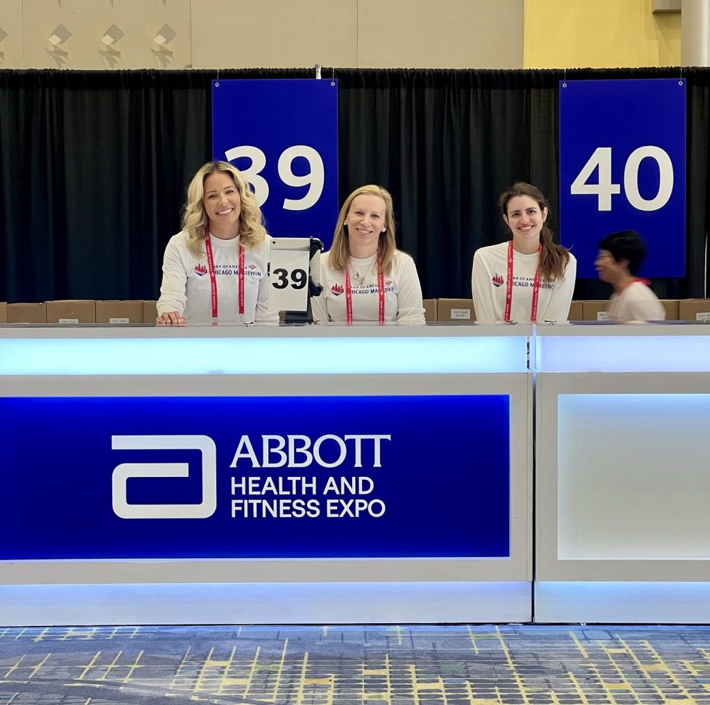 Three women smiling behind a registration desk at the Abbott Health and Fitness Expo, with a large event sign displaying booth numbers 39 and 40 in the background.