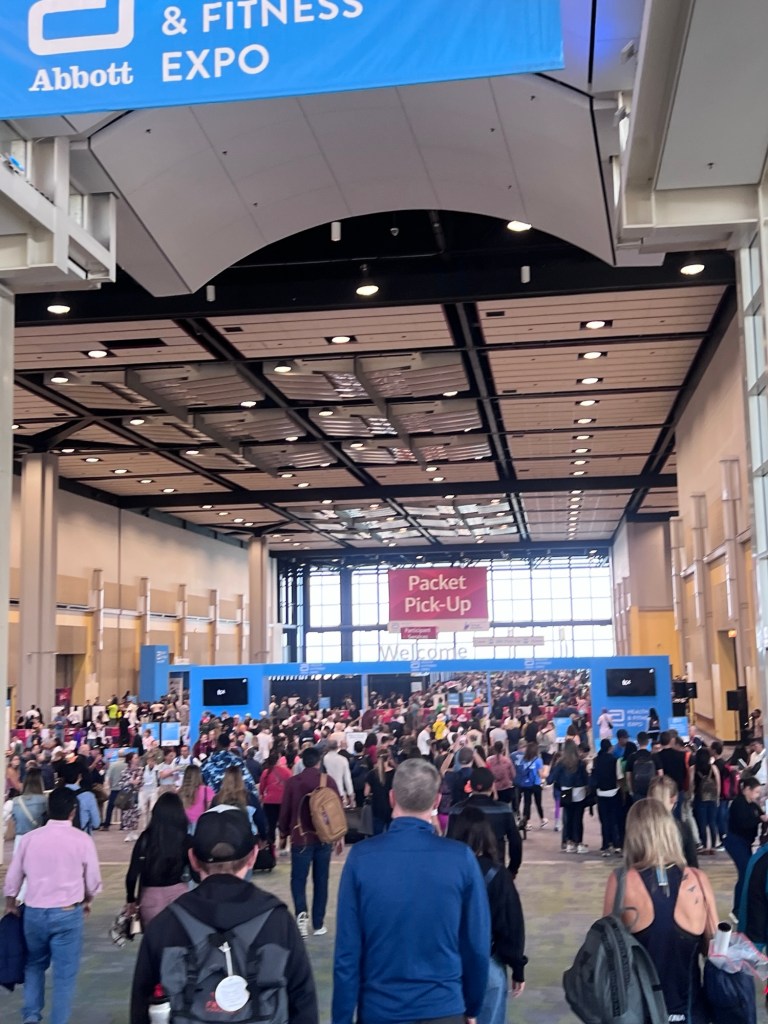 Crowd of attendees entering a convention center for the Abbott Health & Fitness Expo, with a sign for Packet Pick-Up in the background.